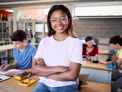 College student in safety glasses standing at a desk with three students in the background.