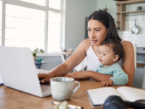 A young mother student holding an infant while sitting at a computer.