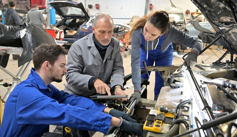 An instructor and two students looking under the hood of a vehicle.